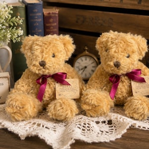 May include: Two identical light brown plush teddy bears with shaggy fur, each wearing a dark magenta satin ribbon tied in a bow around the neck. Each bear has a beige paper tag attached that reads Bears From The Past in black text.