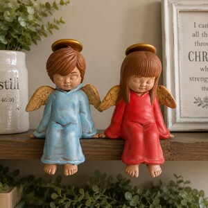 May include: Two angel figurines with closed eyes and gold halos. One figure wears a light blue robe with brown hair, and the other wears a red robe with brown hair. Both have textured gold wings and are sitting with their feet dangling downward.