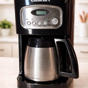 May include: Black Cuisinart coffee maker with a stainless steel carafe. The control panel features a digital clock and buttons for cleaning, brewing, and programming. The carafe is designed to keep coffee warm.