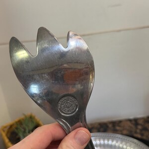 May include: A silver-toned metal salad server with a three-pronged fork head. The surface is reflective and shows signs of use. A circular stamp is visible near the base of the head, featuring the text "WMF" and "CROMARGAN" in a circular pattern.