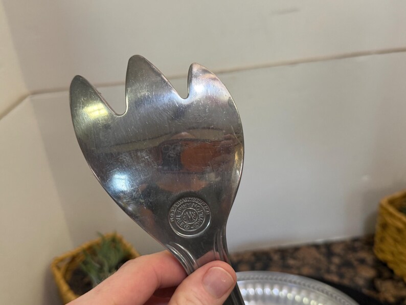 May include: A silver-toned metal salad server with a three-pronged fork head. The surface is reflective and shows signs of use. A circular stamp is visible near the base of the head, featuring the text "WMF" and "CROMARGAN" in a circular pattern.