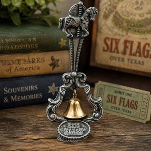 May include: A silver-toned metal souvenir bell featuring a carousel horse at the top and an ornate frame. A small gold-toned bell hangs in the center. The base is an oval plaque with the text SIX FLAGS embossed on the front surface of the metal.