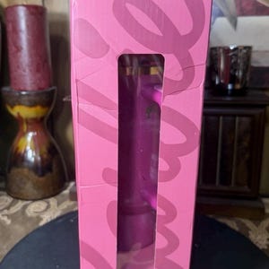 May include: A pink rectangular box with a large rectangular cutout revealing a purple object inside. The box has a design in a darker shade of pink. The box is standing upright.