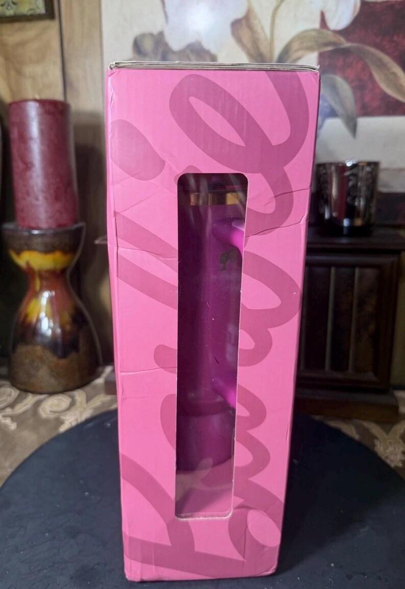 May include: A pink rectangular box with a large rectangular cutout revealing a purple object inside. The box has a design in a darker shade of pink. The box is standing upright.