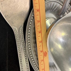 May include: A silver-toned metal serving spoon and a gravy boat with an attached underplate, both featuring a beaded rim design. A wooden ruler is placed between the items, showing a measurement scale from 1 to 9 inches in length.