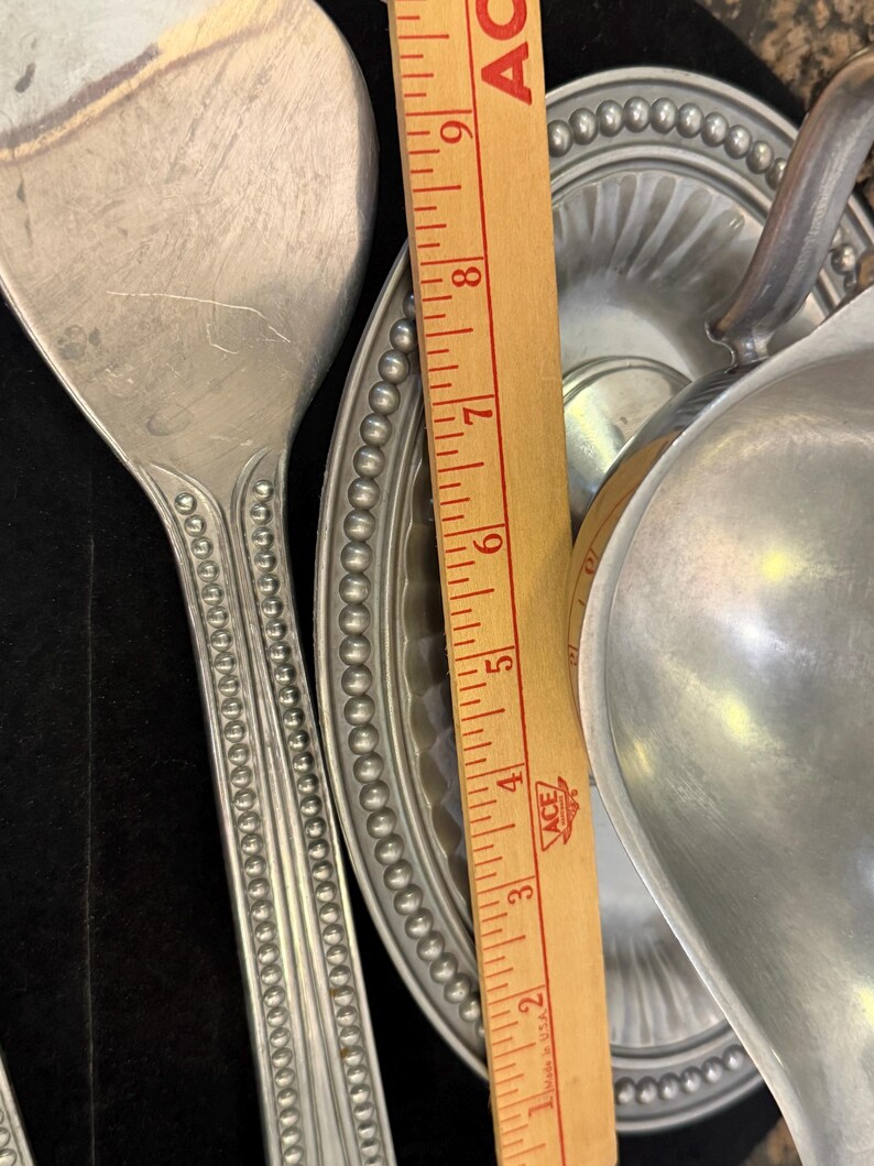May include: A silver-toned metal serving spoon and a gravy boat with an attached underplate, both featuring a beaded rim design. A wooden ruler is placed between the items, showing a measurement scale from 1 to 9 inches in length.