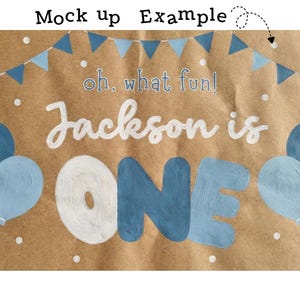 May include: A brown paper backdrop with the text "oh, what fun! Jackson is ONE" in white and blue. The design includes blue balloons and a banner, perfect for a first birthday celebration.
