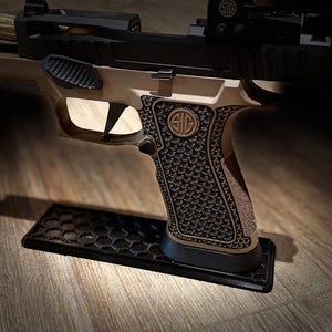 Pode incluir: Uma pistola bege e preta com punho texturizado, com o logotipo SIG. A arma de fogo repousa sobre um suporte preto com padrão de favo de mel. A arma tem um slide preto e uma mira de ponto vermelho.