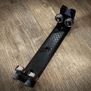 May include: A black, 3D-printed linear rail carriage with four small, silver ball bearings. The carriage has a textured surface and is designed to slide along a track. The item is shown on a wooden surface.