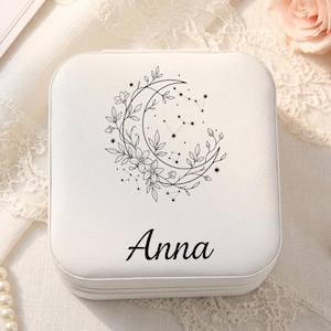 May include: A square white jewelry box featuring a black line art illustration of a crescent moon adorned with flowers and star constellations. The name Anna is printed in black script lettering on the lower portion of the lid surface.