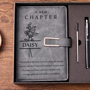 May include: A gray notebook with a rose gold clasp, featuring the text A NEW CHAPTER, DAISY, and a floral graphic. Below, a quote is printed. The notebook is inside a dark gray gift box alongside a black pen and a thin metal refill cartridge.