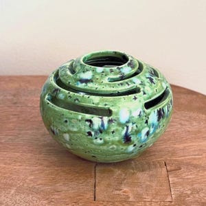 May include: A round, green ceramic decorative object with a spiral design. The object has a glossy finish with blue and black speckles. It sits on a wooden surface. The object has a series of horizontal openings.