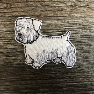 May include: White embroidered patch of a small dog with black eyes and a black nose. The dog is standing on all four legs with its tail curled up.