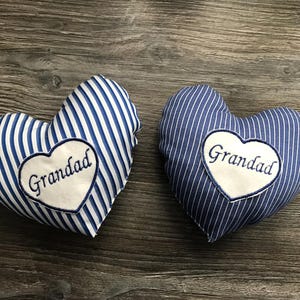 May include: Two heart-shaped cushions with blue and white stripes. Each cushion features a white heart-shaped patch embroidered with the word "Grandad". They are displayed on a wooden surface.