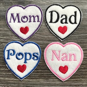 Op de afbeelding: Vier witte hartvormige stoffen patches met geborduurde randen en tekst. De patch linksboven heeft paarse tekst met Mom, rechtsboven zwarte tekst met Dad, linksonder blauwe tekst met Pops en rechtsonder roze tekst met Nan. Elke patch heeft een klein rood hartje onder de tekst.