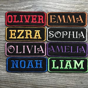 May include: Rectangular embroidered patches with names: OLIVER, EMMA, EZRA, SOPHIA, OLIVIA, AMELIA, NOAH, and LIAM. Each patch features a black background with colorful embroidered lettering and borders in various colors like red, orange, yellow, white, pink, purple, blue, and green.