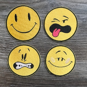 May include: Four yellow embroidered patches with different facial expressions. One patch has a smiling face, another has a face with its tongue sticking out, a third has a worried expression, and the last has a winking face.