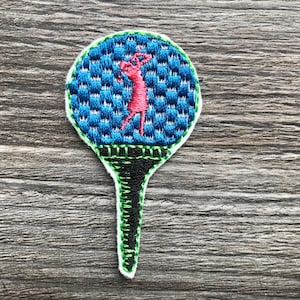 Pode incluir: Etiqueta de bola de golfe bordada com um design azul e verde. Uma figura rosa é representada balançando um taco de golfe. A etiqueta está presa a um tee de golfe verde e preto.