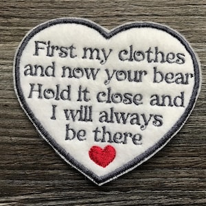 May include: A heart-shaped white fabric patch with a dark grey embroidered border and dark grey text that reads: First my clothes and now your bear Hold it close and I will always be there. A small red embroidered heart sits below the text.