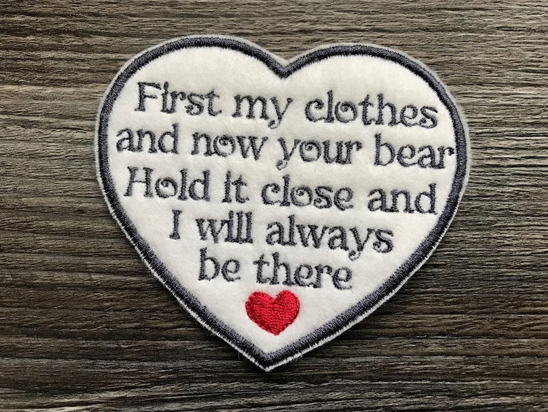 May include: A heart-shaped white fabric patch with a dark grey embroidered border and dark grey text that reads: First my clothes and now your bear Hold it close and I will always be there. A small red embroidered heart sits below the text.