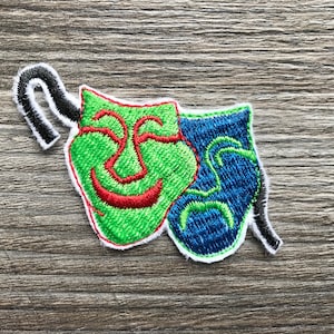 Può includere: Un toppa ricamata che raffigura due maschere teatrali, una sorridente e l'altra accigliata. Le masche sono di colore verde e blu e sono collegate da una linea curva nera.