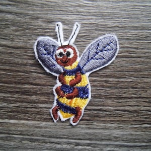 Puede incluir: Una abeja bordada de color amarillo y marrón con rayas azules y una cara sonriente. La abeja tiene dos antenas blancas y alas grises.