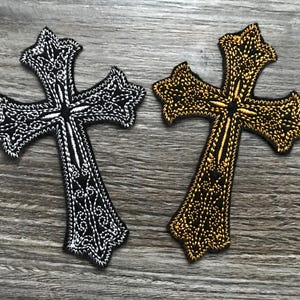 May include: Two embroidered cross patches. One is black with white detailing, the other is gold with black detailing. Both have ornate, decorative patterns and are set against a wood grain background. These patches are suitable for clothing or accessories.