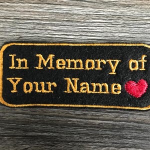 May include: A rectangular black embroidered patch with a gold border. The text "In Memory of Your Name" is stitched in gold thread, accompanied by a small, solid red heart icon positioned at the end of the second line of text.