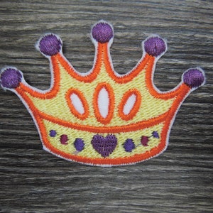 Puede incluir: Una corona bordada de color amarillo con ribete naranja y detalles morados. La corona tiene tres puntas y un corazón en el centro.