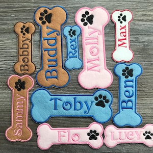 Könnte beinhalten: Eine Sammlung farbenfroher, knochenförmiger Namensschilder für Haustiere. Jedes Schild zeigt einen Pfotenabdruck und einen Namen, der in einer Kontrastfarbe gestickt ist. Namen sind Bobby, Buddy, Rex, Molly, Max, Sammy, Toby, Flo, Ben und Lucy.