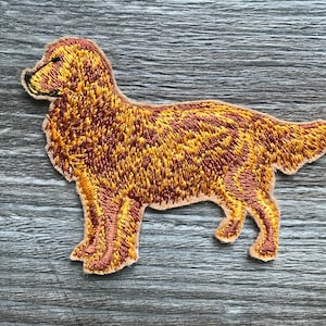 Może przedstawiać: Naszywka haftowana przedstawiająca stojącego golden retrievera z głową zwróconą w bok. Naszywka wykonana jest z brązowej i złotej nici na beżowym materiale.