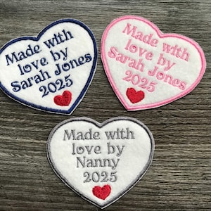 May include: Three heart-shaped embroidered patches. Each patch has a white centre with text that reads "Made with love by" followed by a name and the year 2025. A red heart is at the bottom. The borders are navy, pink, and grey.
