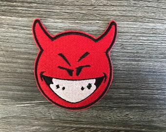 Könnte beinhalten: Rotes Teufelskopf mit einem verschmitzten Grinsen und Hörnern. Das Gesicht ist mit schwarzem Faden auf rotem Stoffgrund gestickt.