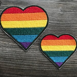 Peut inclure: Deux écussons brodés en forme de cœur. Chaque écusson présente un motif arc-en-ciel avec des rayures rouges, orange, jaunes, vertes, bleues et violettes. Les écussons ont une bordure noire et sont posés sur un fond en bois.