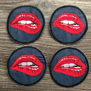 May include: Four round denim patches with embroidered red lips. Each patch features a pair of red lips with white teeth, outlined in black thread. The patches are arranged in a square formation.