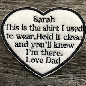 Pode incluir: Um emblema de tecido branco em forma de coração com uma borda bordada em preto. O emblema apresenta um texto bordado em preto que diz: Sarah This is the shirt I used to wear. Hold it close and you'll know I'm there. Love Dad.