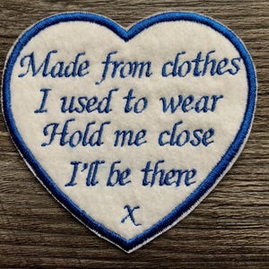 May include: A heart-shaped white fabric patch with a blue embroidered border and blue text that reads: Made from clothes I used to wear Hold me close I'll be there x. The patch features a clean, stitched edge and is designed for attachment to textiles.