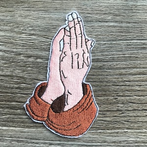 Peut inclure: Patch brodé de deux mains jointes en prière, les mains étant de couleur rose clair et les manches de couleur marron.