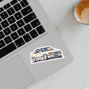 Op de afbeelding: Een witte pixel art auto sticker met een zwarte omtrek is geplaatst op een laptop. De auto heeft een coupé-ontwerp met een spoiler en zilveren wielen. Een kopje koffie is rechtsboven zichtbaar.