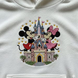 May include: White fleece hoodie featuring an embroidered design of Mickey Mouse and Minnie Mouse in front of a pink and blue castle. The design includes gold stars and trees, creating a whimsical theme.