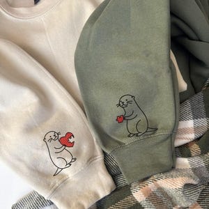 May include: Cream and olive green sweatshirts with embroidered otter designs. One otter holds a red heart, the other a red gift. The sweatshirts are folded over a plaid blanket.