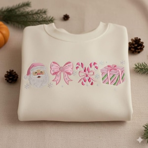May include: Cream-colored sweatshirt with embroidered Christmas-themed designs. The design includes Santa Claus, a pink bow, candy canes, and a wrapped gift. The embroidery is in shades of pink and white, creating a festive and cheerful look.
