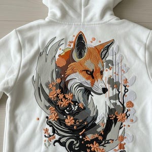 Könnte beinhalten: Weißer Kapuzenpullover mit einem gestickten Fuchsmotiv. Der Fuchs ist mit orangefarbenem, braunem und grauem Fell dargestellt, umgeben von floralen Akzenten. Die Stickerei ist detailliert und hat eine Kapuze.