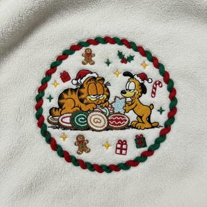 Puede incluir: Parche bordado con Garfield y un perro, ambos con gorros de Papá Noel, rodeados de galletas y regalos navideños. El diseño está enmarcado por un borde trenzado rojo, verde y blanco, con estrellas y acentos de acebo. Sobre un fondo blanco.