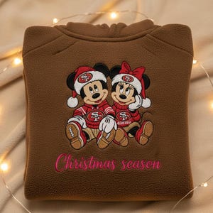 Peut inclure: Sweat-shirt en polaire marron avec une broderie de Mickey et Minnie Mouse vêtus de tenues de football des 49ers. Le texte "Christmas season" est en rose. Les personnages portent des bonnets de Père Noël et tiennent des ballons de football.