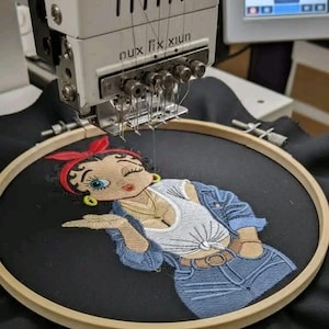 Op de afbeelding: Een borduurmachine borduurt een Betty Boop-ontwerp op zwarte stof. Het ontwerp toont Betty Boop in een spijkerjasje, witte top en rode haarband. De machine is wit met de tekst "nux fix xiun". Een scherm toont het ontwerp.