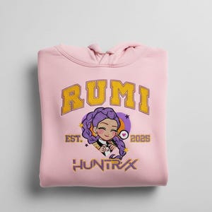 Op de afbeelding: Een lichtroze hoodie met de tekst "RUMI" in gouden letters. Daaronder een cartoon illustratie van een meisje met paars haar. De tekst "EST. 2026" en "HUNTRX" zijn ook zichtbaar.