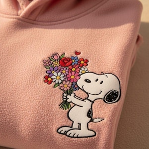 May include: Pale pink fleece hoodie with an embroidered Snoopy design holding a bouquet of flowers. The flowers are a mix of colours, including red and white. The hoodie has a hood and a soft texture.