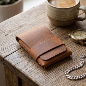 May include: A brown leather wallet with a card slot is on a wooden table, next to a cup of coffee, a watch, and a silver chain bracelet. A succulent plant in a ceramic pot and a book are also on the table.