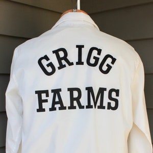 May include: White jacket with long sleeves and a black embroidered logo that says "GRIGG FARMS" on the back.